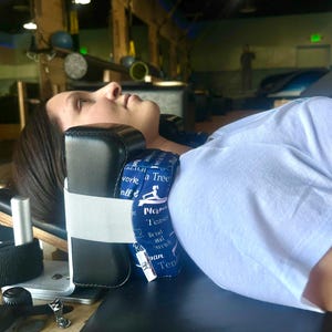 May include: A black and white neck support pillow with a white elastic strap. The pillow has blue fabric with white text and a yoga graphic. The person is lying on a black surface.