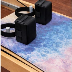 May include: Black exercise blocks with straps and a colorful mat with a blue, purple, and pink ocean-like design. The blocks are rectangular and appear to be made of leather or a similar material. The equipment is on a wooden frame.
