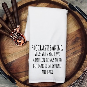 May include: A white kitchen towel featuring the black printed text: PROCRASTIBAKING VERB: WHEN YOU HAVE A MILLION THINGS TO DO BUT IGNORE EVERYTHING AND BAKE. Beside the towel are copper measuring spoons with dark wooden handles.