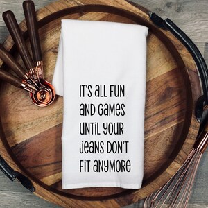 May include: A white kitchen towel with the text "IT'S ALL FUN AND GAMES UNTIL YOUR JEANS DON'T FIT ANYMORE" is displayed on a wooden tray. Copper measuring spoons and a whisk are also on the tray.