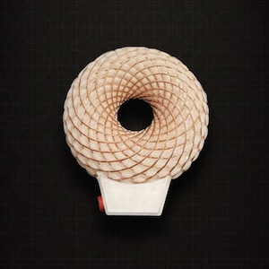 Pode incluir: Um objeto único em forma de donut, de cor clara, com um buraco central, sobre uma base retangular branca com um detalhe vermelho. O objeto tem uma textura espiralada e tecida. O fundo é uma superfície escura e padronizada.