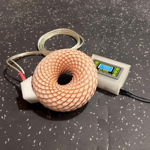 Pode incluir: Um objeto complexo em forma de donut com um padrão tecido castanho claro. Está conectado a um componente retangular branco e a uma unidade de exibição digital com o texto "ZK-PP2K High Power PWM Driver". Um cabo prateado também está anexado.