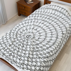 Pode incluir: Uma colcha branca com um padrão geométrico preto de círculos e números interligados. O design apresenta um "9" central rodeado por anéis concêntricos de números e linhas. A colcha está sobre uma estrutura de cama de madeira.