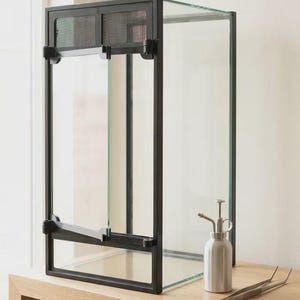 May include: A vertical glass terrarium with a black metal frame and a front-opening door featuring a mesh ventilation panel at the top. Beside the enclosure sits a small silver metal spray bottle and a pair of long, thin metal tweezers.