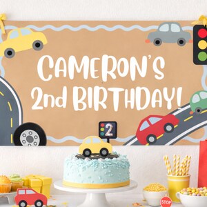 May include: A birthday party scene with a banner that reads "Cameron's 2nd Birthday!" featuring car illustrations. A light blue cake with a yellow base is topped with a yellow car. Other decorations include cupcakes, candies, and a red gift bag.