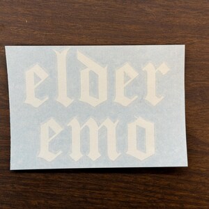 May include: White sticker with the words "elder emo" in a gothic font. The sticker is on a light blue background, set against a dark brown wooden surface. The text is the focal point of the image.