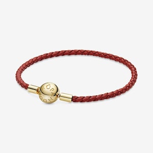 May include: A red braided leather bracelet with a gold-colored clasp. The clasp is a round sphere with the brand name engraved on it. The bracelet is a stylish accessory.