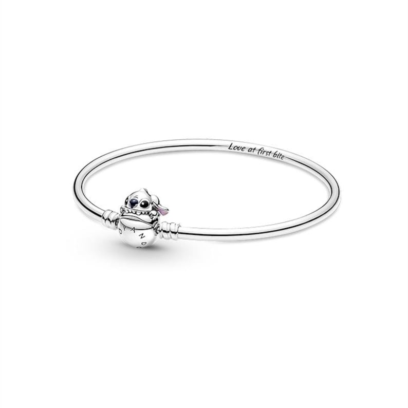 May include: A silver bracelet featuring a charm of a cartoon character with blue and pink accents. The charm is attached to a thin, polished silver band. The bracelet has the inscription "Love at first bite".