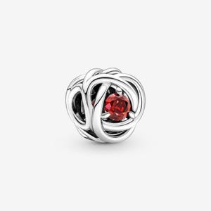 Puede incluir: Un charm de plata con un diseño tejido, con una gema roja vibrante en el centro. El charm es redondo y tiene un acabado pulido, adecuado para pulseras o collares. La gema es de color rojo intenso.