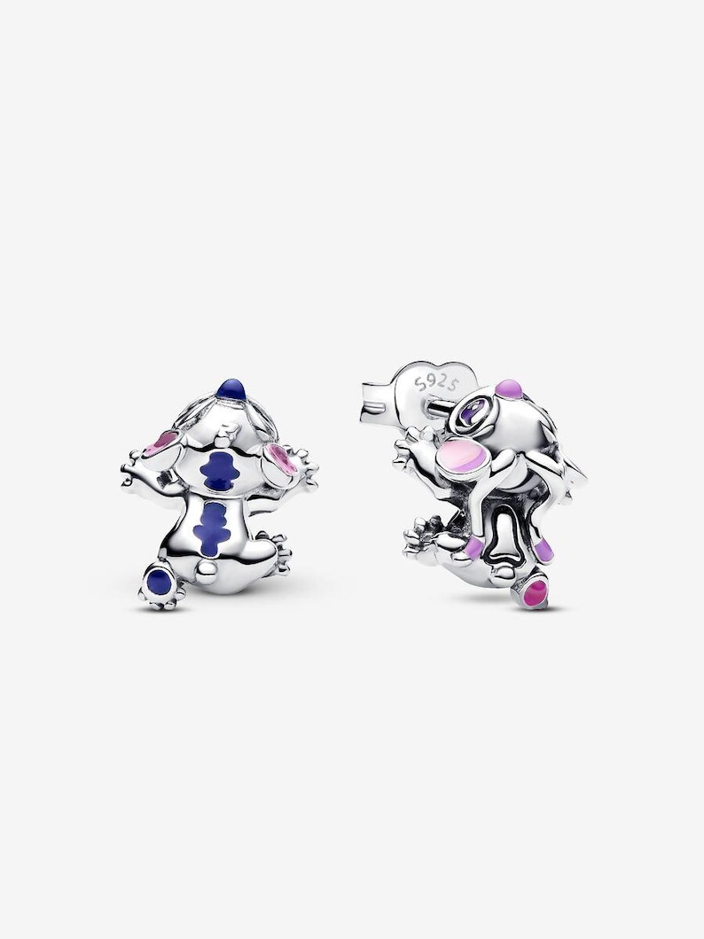 May include: A pair of silver-toned stud earrings featuring a cartoon character design. The character has blue, pink, and purple accents. The earrings are detailed with small arms and legs, and a post backing.