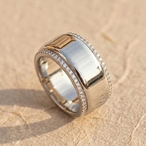 Puede incluir: Un anillo plateado con una banda ancha y una fila de pequeñas piedras transparentes. El anillo tiene una superficie pulida y reflectante y un acento dorado en el borde superior. El anillo está sobre un fondo neutro y texturizado.