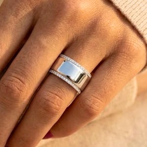 Puede incluir: Un anillo plateado con un centro rectangular y una banda de pequeñas piedras brillantes. El anillo está en un dedo, con un suéter de color claro en el fondo. El anillo es una pieza llamativa.