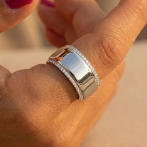 Puede incluir: Un anillo plateado con un centro rectangular y una banda de pequeñas piedras preciosas transparentes. El anillo está en un dedo con esmalte de uñas rojo. El anillo tiene una superficie pulida y reflectante.