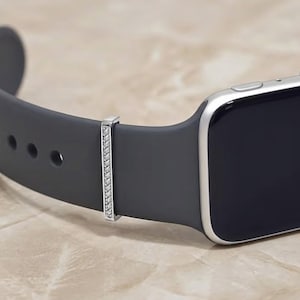 Puede incluir: Primer plano de una correa de reloj inteligente gris oscuro con una banda decorativa plateada. La banda decorativa es rectangular e incrustada con pequeñas piedras preciosas transparentes. La esfera del reloj es negra con un borde plateado.