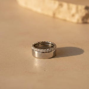 Könnte beinhalten: Zwei Silberringe, einer übereinander gestapelt. Der obere Ring ist mit einer Reihe kleiner, klarer Steine besetzt. Der untere Ring ist ein schlichtes, poliertes Band. Die Ringe sind vor einem neutralen Hintergrund platziert.