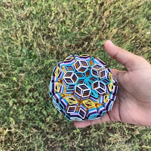 Puede incluir: Una esfera geométrica multicolor compuesta por formas entrelazadas en tonos azules, morados, amarillos y blancos. La esfera se sostiene en una mano sobre un fondo de hierba verde.