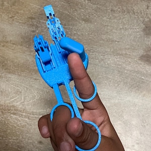 May include: A light blue 3D-printed prosthetic finger attachment. The device has articulated joints and finger rings for attachment. The device is designed to assist with gripping and manipulation.