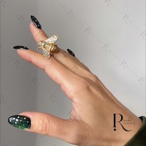 Puede incluir: Un anillo de abeja dorado con detalles de diamantes. El anillo presenta un diseño detallado de una abeja con las alas extendidas. El anillo se lleva en un dedo, mostrando la intrincada artesanía y los detalles brillantes de la joya.