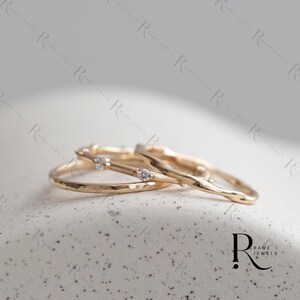 Puede incluir: Tres anillos de oro con texturas y diseños únicos. Uno presenta un acabado martillado, mientras que los otros tienen un aspecto liso y orgánico. Dos anillos están adornados con pequeñas piedras preciosas transparentes. Los anillos se exhiben sobre una superficie moteada.