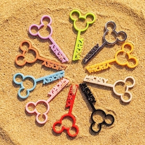 May include: A collection of ten plastic keys arranged in a circle, each featuring a Mickey Mouse head silhouette at the top and the word MICKEY embossed on the shaft. The keys are colored in pink, lime green, dark gray, orange, yellow, light blue, white, red, and black.