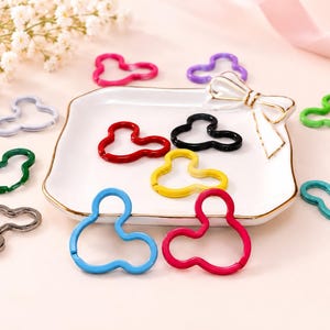 May include: A collection of metal spring gate rings in various colors including red, blue, yellow, black, green, and purple. These circular connectors feature a three-lobed shape and are arranged around a white square dish with a decorative bow.