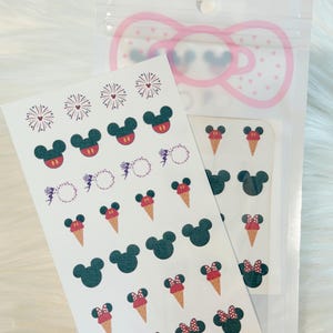 May include: A sheet of adhesive stickers featuring various designs including dark green mouse-shaped icons, mouse-shaped icons with red shorts, mouse-shaped icons with red polka-dot bows, ice cream cones, fireworks, and fairy silhouettes in a clear bag.