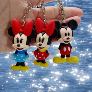 May include: Three character keychains featuring two figures with red bows, one wearing a blue polka-dot dress and the other a red polka-dot dress, alongside one figure in red shorts and yellow shoes, all attached to silver-toned metal key rings.