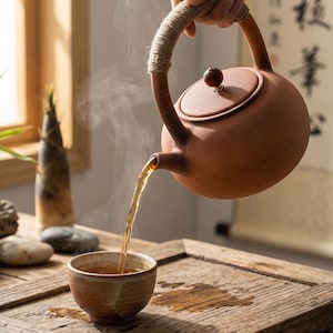 Könnte beinhalten: Eine braune Keramik-Teekanne mit einem griffumwickelten Seil gießt Tee in eine kleine, passende Tasse. Dampf steigt vom Tee auf, was darauf hindeutet, dass er heiß ist. Die Teekanne und die Tasse stehen auf einem Holztablett und schaffen eine ruhige Teestunde.