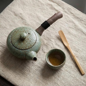 May include: A celadon green ceramic teapot with a wooden handle and a small spout. A small ceramic cup and a wooden spoon are next to the teapot. The teapot has a textured surface and a black band around the handle.