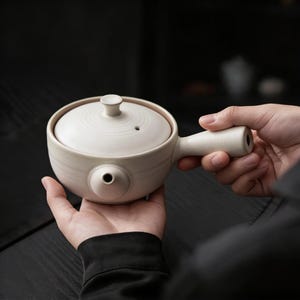 May include: A cream-colored ceramic teapot with a round body, a lid, and a handle. The teapot has a small spout and a circular opening on the side. The lid has a small knob on top. The teapot is held in two hands.