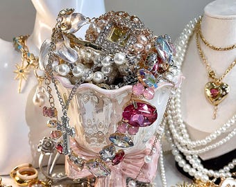 May include: A collection of jewelry, including necklaces, rings, and earrings, displayed on white busts and a pink pedestal. The jewelry features pearls, crystals, and various gemstones in shades of pink, gold, and silver. An eye-shaped earring is visible.
