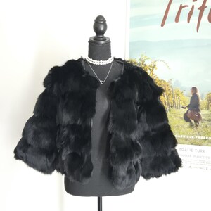 May include: A black fur jacket with a layered, horizontal panel design and three-quarter length sleeves. The jacket features an open front, a collarless neckline, and a soft, dense texture, displayed on a black mannequin with a pearl necklace.