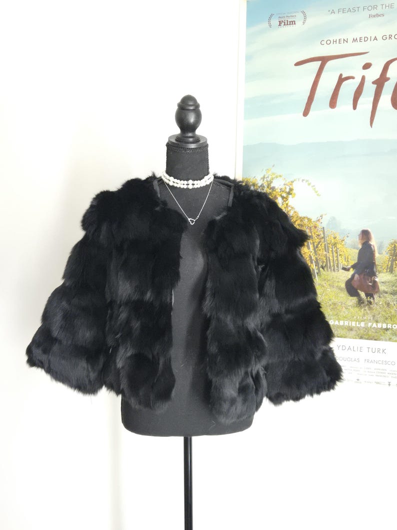 May include: A black fur jacket with a layered, horizontal panel design and three-quarter length sleeves. The jacket features an open front, a collarless neckline, and a soft, dense texture, displayed on a black mannequin with a pearl necklace.