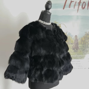 May include: A black, cropped, long-sleeved jacket made of layered, textured fur. The jacket features a round neckline and is displayed on a black mannequin torso, which is adorned with a double-strand white pearl necklace around the neck.