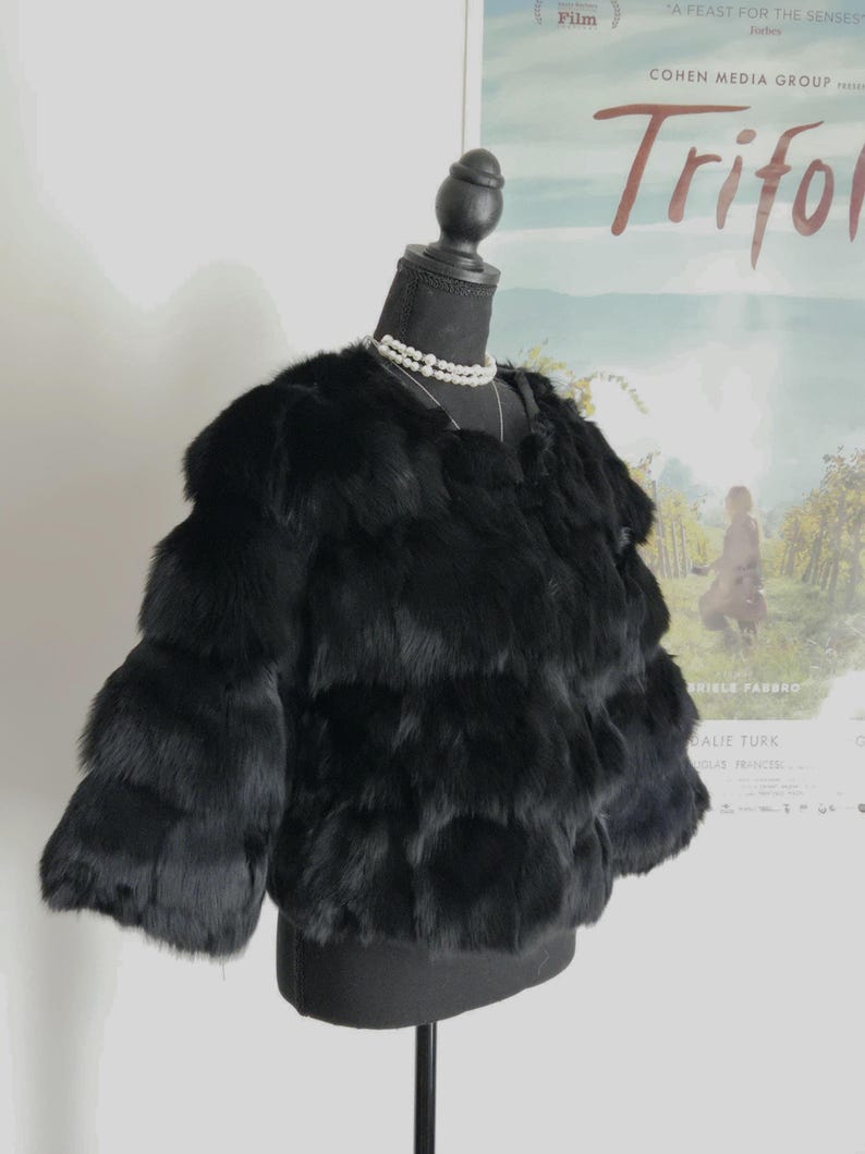 May include: A black, cropped, long-sleeved jacket made of layered, textured fur. The jacket features a round neckline and is displayed on a black mannequin torso, which is adorned with a double-strand white pearl necklace around the neck.