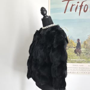 May include: A black, long-sleeved, textured fur jacket displayed on a black mannequin torso. The jacket features a layered, shaggy appearance with a round neckline. A double-strand white pearl necklace is worn around the neck of the mannequin.