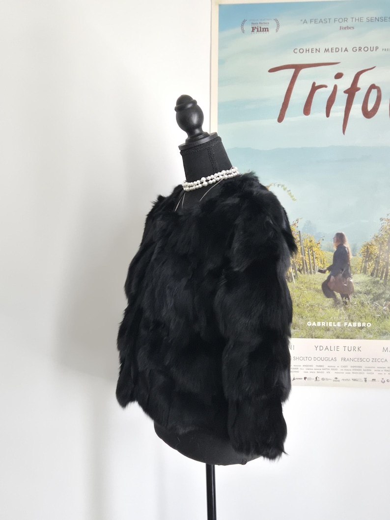 May include: A black, long-sleeved, textured fur jacket displayed on a black mannequin torso. The jacket features a layered, shaggy appearance with a round neckline. A double-strand white pearl necklace is worn around the neck of the mannequin.