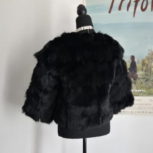 May include: A black cropped jacket with a textured, layered appearance, displayed on a mannequin. The garment features short sleeves and a rounded neckline, showcasing a dense, soft-looking surface texture throughout the entire piece.