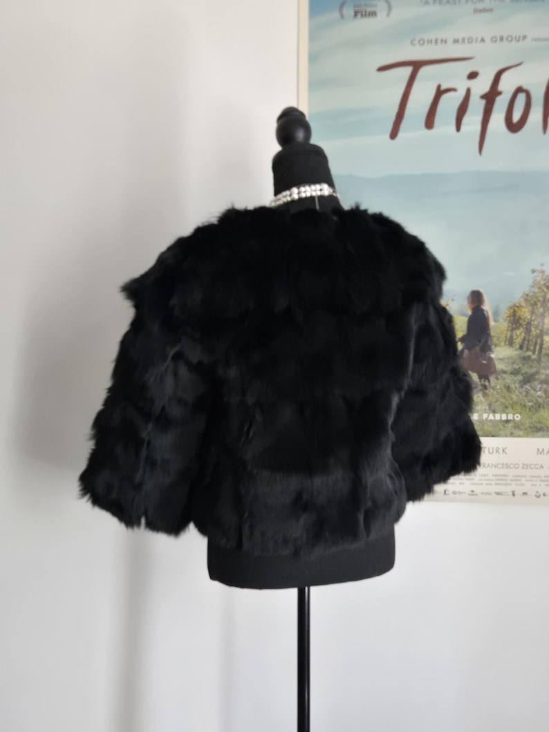 May include: A black cropped jacket with a textured, layered appearance, displayed on a mannequin. The garment features short sleeves and a rounded neckline, showcasing a dense, soft-looking surface texture throughout the entire piece.