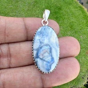 May include: An oval pendant featuring a polished blue and white stone set in a silver-toned metal frame with a scalloped edge. The stone displays marbled patterns of deep blue, light blue, and white, and includes a silver-toned metal bail at the top.