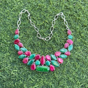 May include: A silver necklace with red and green gemstones. The necklace features teardrop and oval-shaped stones set in a silver frame, connected by a silver chain. The necklace is displayed on a green surface.