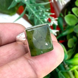 Op de afbeelding: Een zilveren ring met een grote, rechthoekige, olijfgroene edelsteen. De edelsteen heeft een gepolijst oppervlak met subtiele kleurvariaties. De ring wordt in een hand gehouden tegen een achtergrond van groen gebladerte.