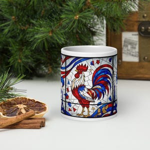 May include: White ceramic mug featuring a stained-glass design of a rooster in red, white, and blue. The rooster stands on a blue and gold base, with the Eiffel Tower in the background. The mug has a black rim and handle.