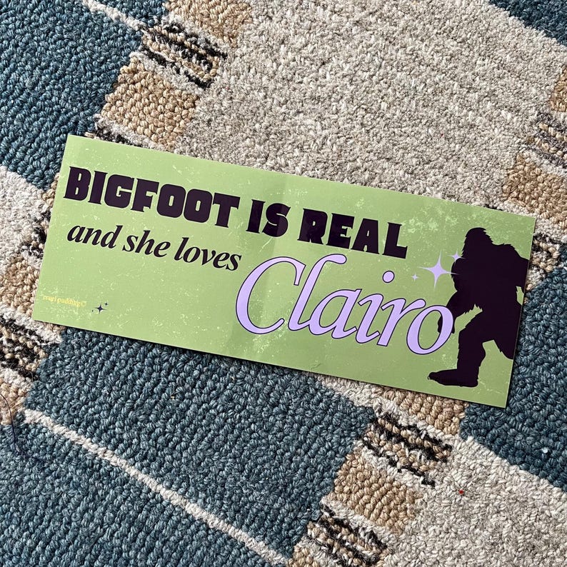 Może przedstawiać: Prostokątna naklejka z jasnozielonym tłem. Tekst "BIGFOOT IS REAL and she loves Clairo" jest w kolorze czarnym i fioletowym. Czarna sylwetka Bigfoota znajduje się po prawej stronie naklejki.