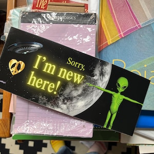 May include: A black rectangular sign with the text "I'm new here!" in yellow neon lettering. The sign features a gold peace sign heart, a UFO, and a green alien with outstretched arms. The background includes a planet and a black space.