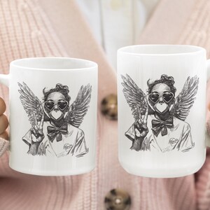Puede incluir: Dos tazas de cer&aacute;mica blanca con una ilustraci&oacute;n en blanco y negro de una persona con alas de &aacute;ngel, gafas de sol en forma de coraz&oacute;n y un signo de la paz. La persona tiene un coraz&oacute;n sobre la boca y una pajarita.