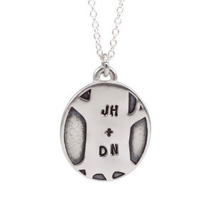 Op de afbeelding: Zilveren ketting met een ronde hanger met de initialen "JH" en "DN", gescheiden door een klein kruis.