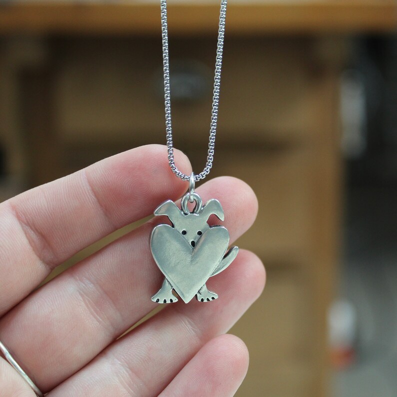 May include: A silver necklace with a pendant shaped like a dog holding a heart in its paws.