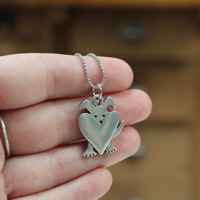 May include: Silver necklace with a charm of a dog holding a heart in its paws.