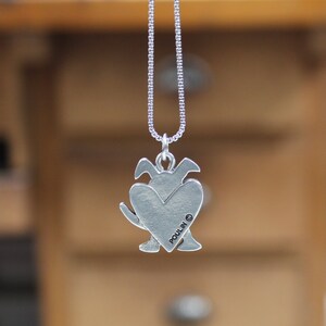 May include: Silver necklace with a heart-shaped pendant featuring a dog silhouette. The back of the pendant is engraved with the word "POULIN" and a copyright symbol.